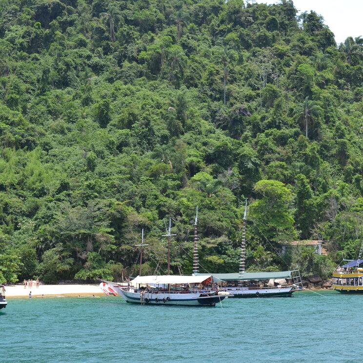 Anlegestelle mit traditionellen Segelschiffen in Paraty, Brasilien, umgeben von üppiger tropischer Vegetation und Strand.