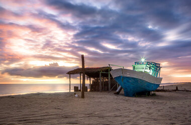 Blau-weißes Holzboot und eine einfache Strohhütte am Sandstrand von Prea bei Sonnenuntergang.