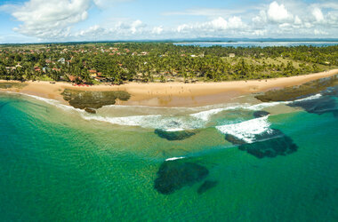 Luftaufnahme der Praia dos Três Coqueiros auf der Halbinsel Maraú. Ein breiter, goldener Sandstrand schmiegt sich an ein türkisblaues Meer mit sichtbaren Korallenriffen unter der Wasseroberfläche. Das Hinterland ist dicht mit Palmen bewachsen, dazwischen blitzen vereinzelte Häuser hervor.