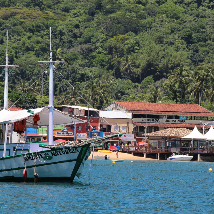 Traditionelles Fischerboot im Wasser vor der Küste von Ilha Grande, Brasilien, mit bunten Geschäften und der Pousada Bela Mar im Hintergrund.