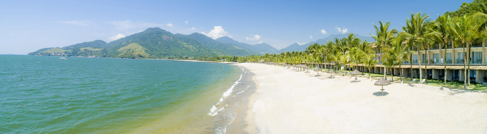 Sandstrand und Hotelgebäude des Portobello Resorts in Angra dos Reis, Brasilien, an türkisblauem Meer vor Bergen.