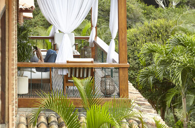 Überdachter Balkon im PortoBay Buzios mit weißen Vorhängen, Holzmöbeln und Blick auf tropische Palmen in Brasilien.