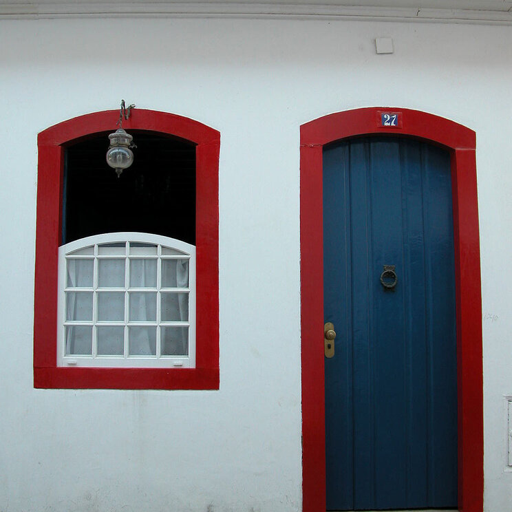 Detailaufnahme einer Wand mit rotem Fensterrahmen und blauer Tür in Paraty, Brasilien, die die charmante koloniale Architektur und die lebhaften Farben der Stadt hervorhebt.