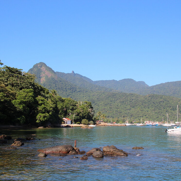 Blick auf die Küste von Ilha Grande, Brasilien, mit sanften Hügeln, üppiger Vegetation und Booten im Wasser, umgeben von einer klaren, blauen Himmel.