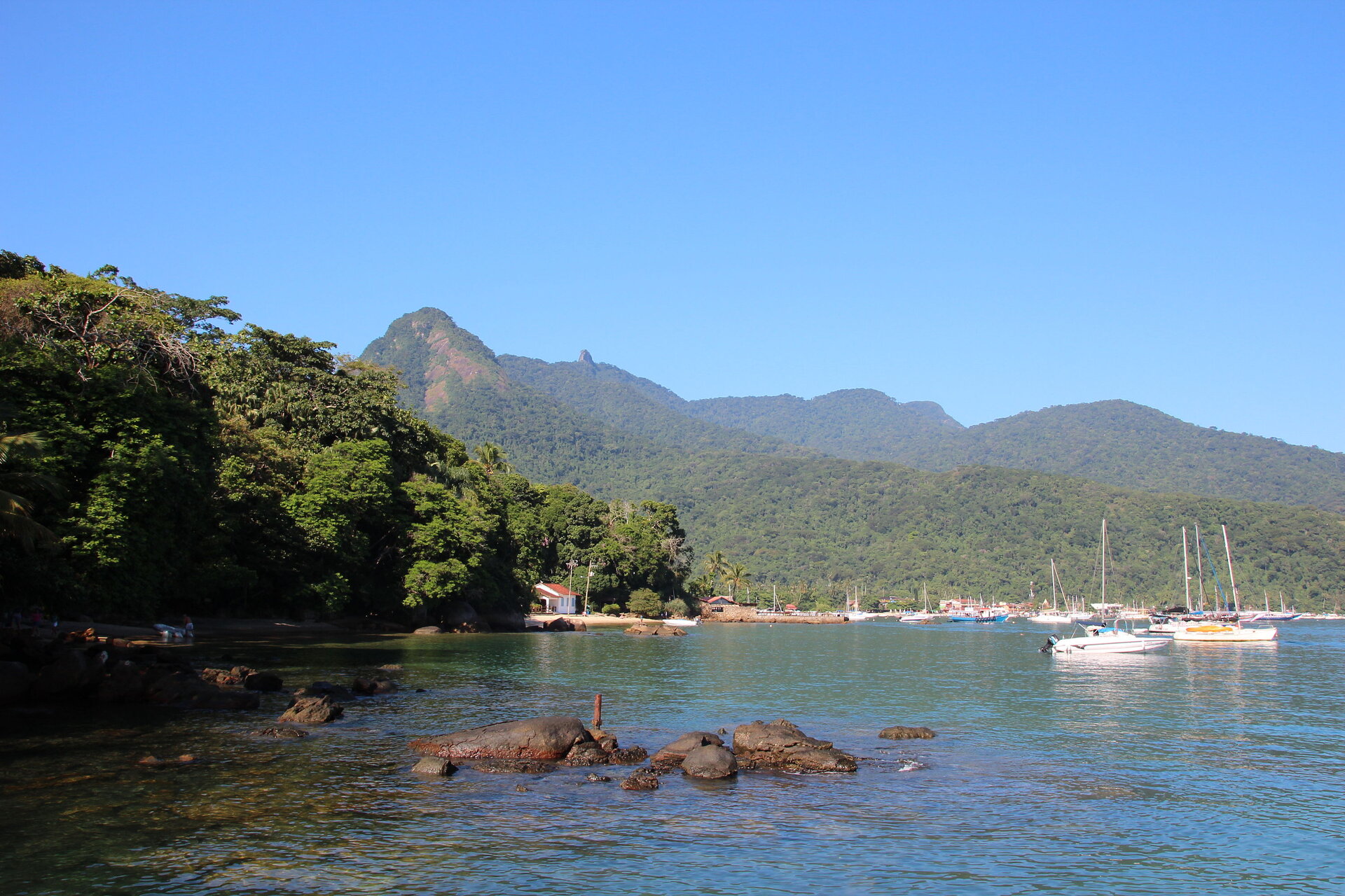 Blick auf die Küste von Ilha Grande, Brasilien, mit sanften Hügeln, üppiger Vegetation und Booten im Wasser, umgeben von einer klaren, blauen Himmel.