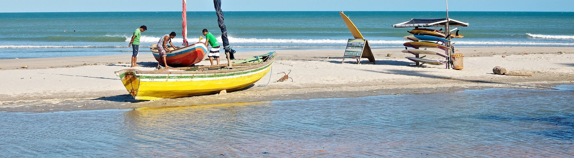 Fischerboote und Surfbretter am Strand von Jericoacoara, mit Einheimischen, die an der Küste aktiv sind, umgeben von klarem Wasser und strahlendem Himmel.