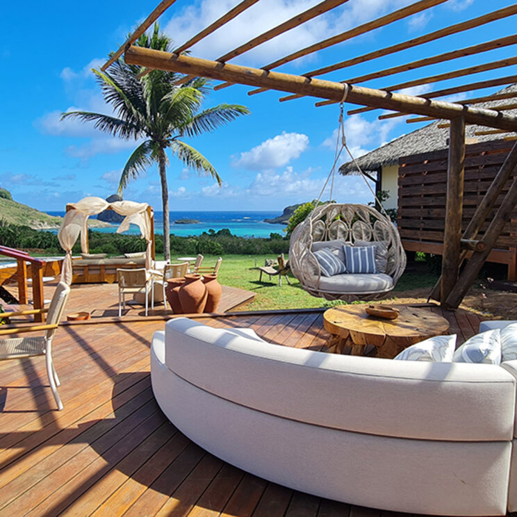 Modernes Holzdeck mit runder Couch, Haengestuhl und Blick auf das Meer unter blauem Himmel.