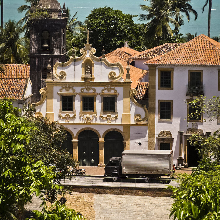 Ein malerischer Blick auf die Küste von Olinda, mit historischen Gebäuden, umgeben von Palmen und dem blauen Meer im Hintergrund.