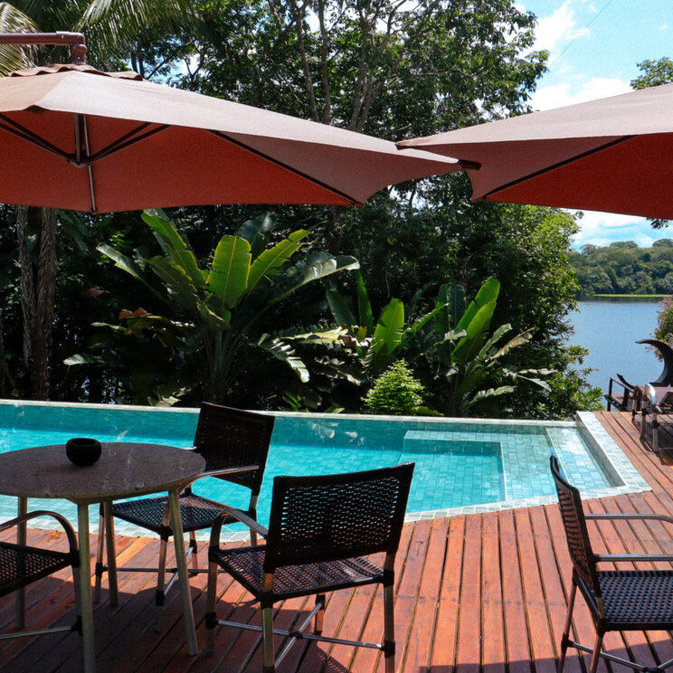 Pool mit Holzterrasse, Sitzgruppen, Sonnenschirmen, tropischen Pflanzen und Blick auf den Fluss bei Manaus in Brasilien.