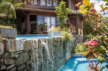 Hotelgebaude aus Holz und Stein mit Pool und integriertem Wasserfall in den Garten.