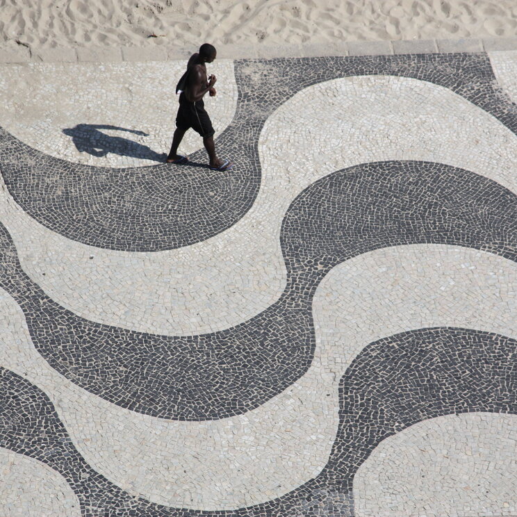 Mann, der über das charakteristische Wellenmuster der Gehwege in Copacabana, Rio de Janeiro, spaziert, mit der lebhaften Atmosphäre und dem Sandstrand im Hintergrund.