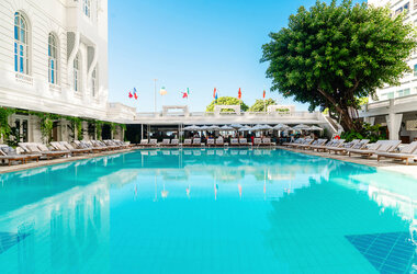 Grosser, hellblauer Hotelpool mit zahlreichen Liegestuehlen unter Flaggen vor weisser Kolonialarchitektur.