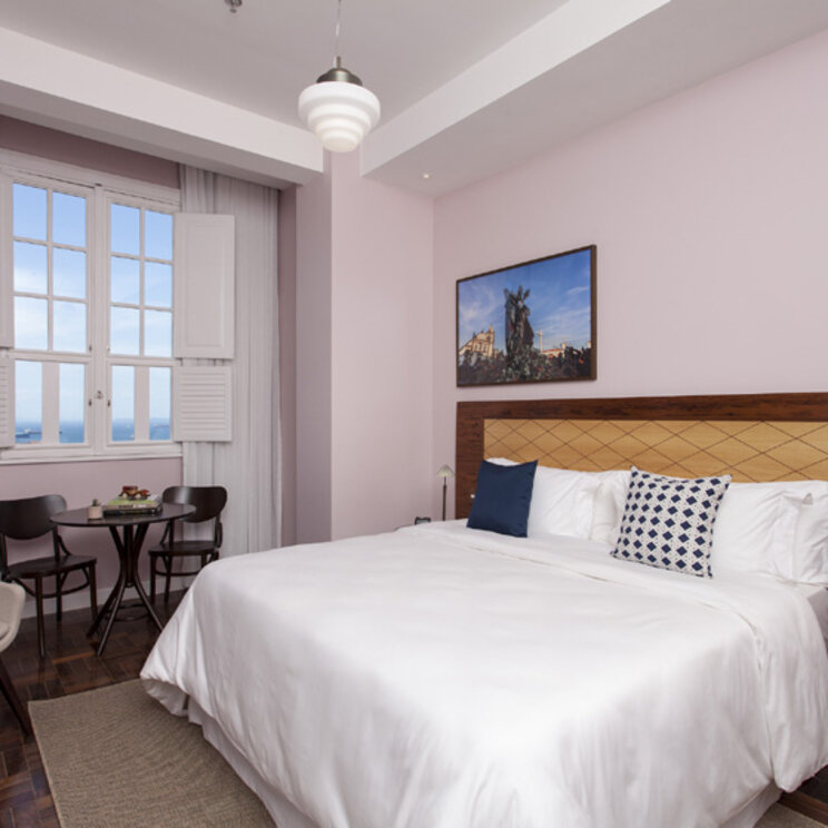 Helles Hotelzimmer mit Doppelbett, Parkettboden und Blick auf das Meer durch weiße Fensterläden in Salvador.