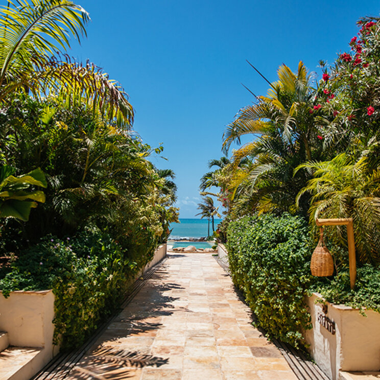 Gepflasterter Gartenweg mit tropischen Pflanzen und Palmen sowie Blick auf das Meer in Jericoacoara, Brasilien.