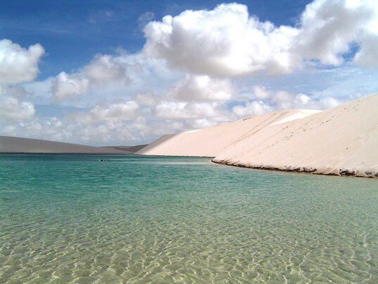 Kristallklares Wasser in einer Lagune des Lençóis Maranhenses Nationalparks, umgeben von weißen Sanddünen und einem strahlend blauen Himmel mit sanften Wolken.