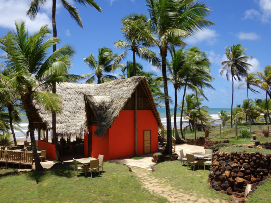 Außenansicht des Hotelrestaurants der Pousada Capitu mit leuchtend oranger Wand und traditionellem Strohdach inmitten einer grünen Palmenlandschaft am Strand.
