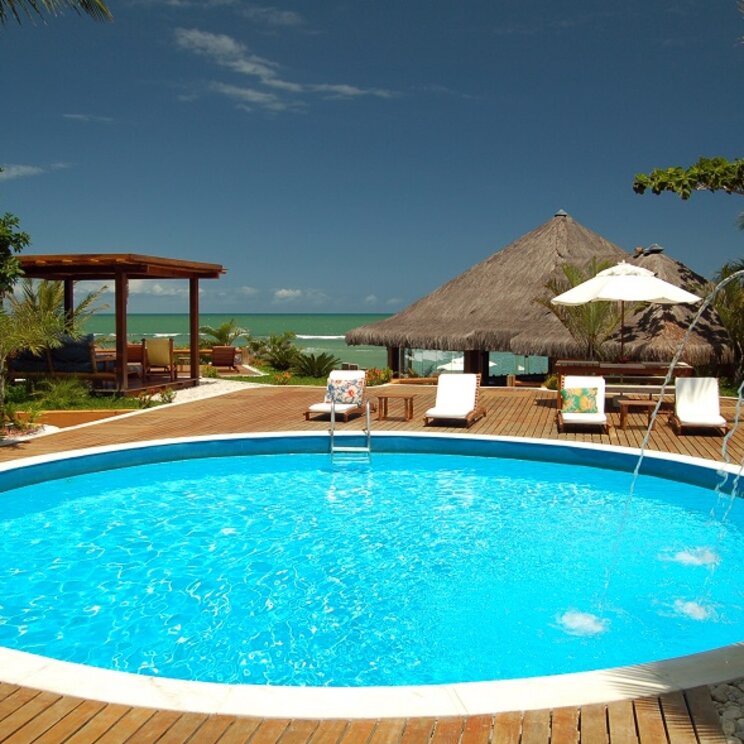 Poolbereich der Pousada Pitinga in Porto Seguro, Brasilien, mit Blick auf das Meer und umgeben von tropischen Hütten und Liegestühlen.