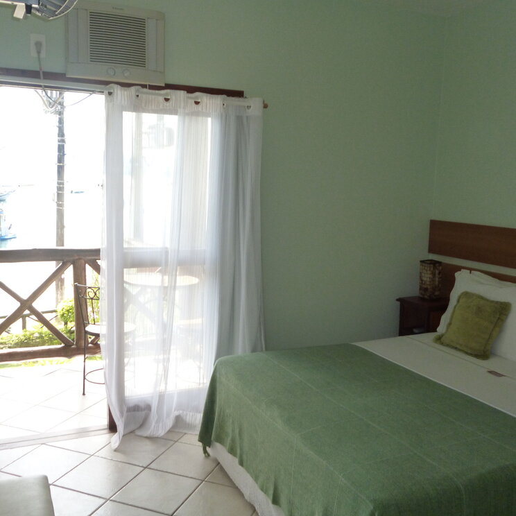 Helles Zimmer im Hotel Pousada Caiçara auf Ilha Grande, mit einem gemütlichen Doppelbett, einer kleinen Terrasse und Blick auf die Umgebung durch geöffnete Vorhänge.