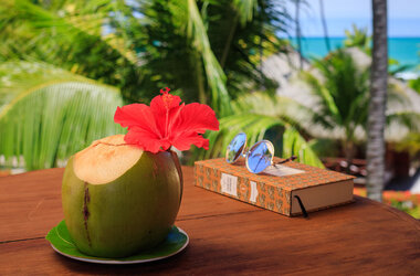 Frische grüne Kokosnuss mit Hibiskusblüte neben Buch und Sonnenbrille auf Holztisch im Nannai Resort, Brasilien.