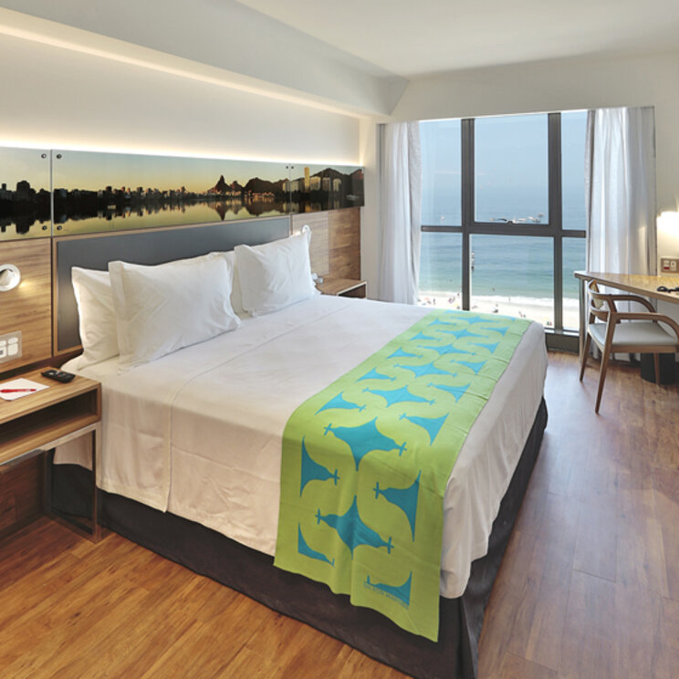 Modernes Hotelzimmer mit Doppelbett, Holzboden und großem Fenster mit Blick auf den Strand in Rio de Janeiro.