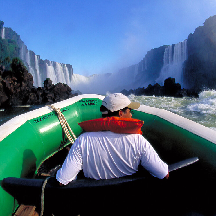 Blick aus einem Schlauchboot auf die Foz do Iguaçu-Wasserfälle, mit einem Mann im Boot, der von den mächtigen Wasserfällen und der Gischt umgeben ist.