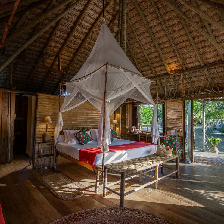 Bungalow-Innenraum mit Reetdach, Doppelbett samt Moskitonetzen und Holz-Sitzbank im Rancho do Peixe in Preá, Brasilien.
