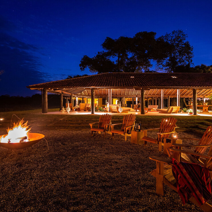 Refúgio Ecológico Caiman bei Nacht mit brennender Feuerschale, Holzsesseln und beleuchteter Lodge.