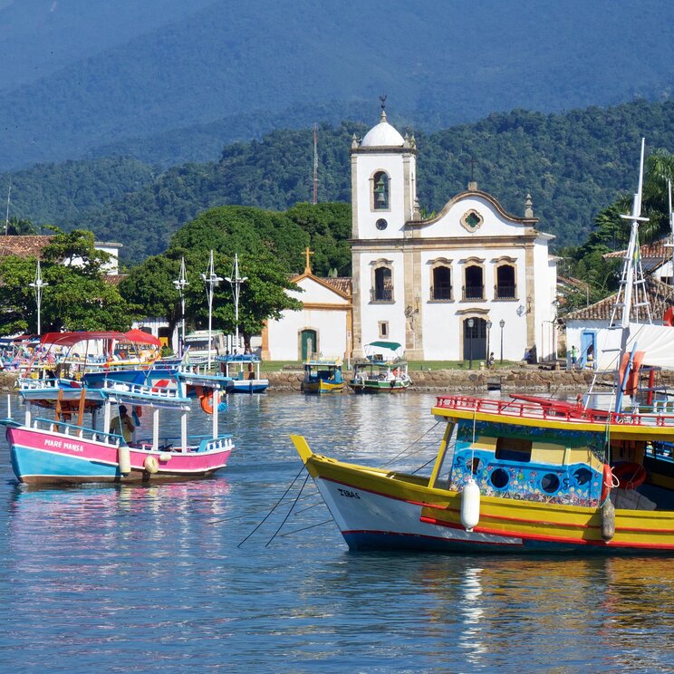 Bunte Holzboote liegen in der Bucht der kolonialen Stadt Paraty, in Brasilien. Im Hintergrund ist eine koloniale Kirche und die Gebirgskette Serra da Bocaina mit dem Atlantischen Regenwald zu sehen.