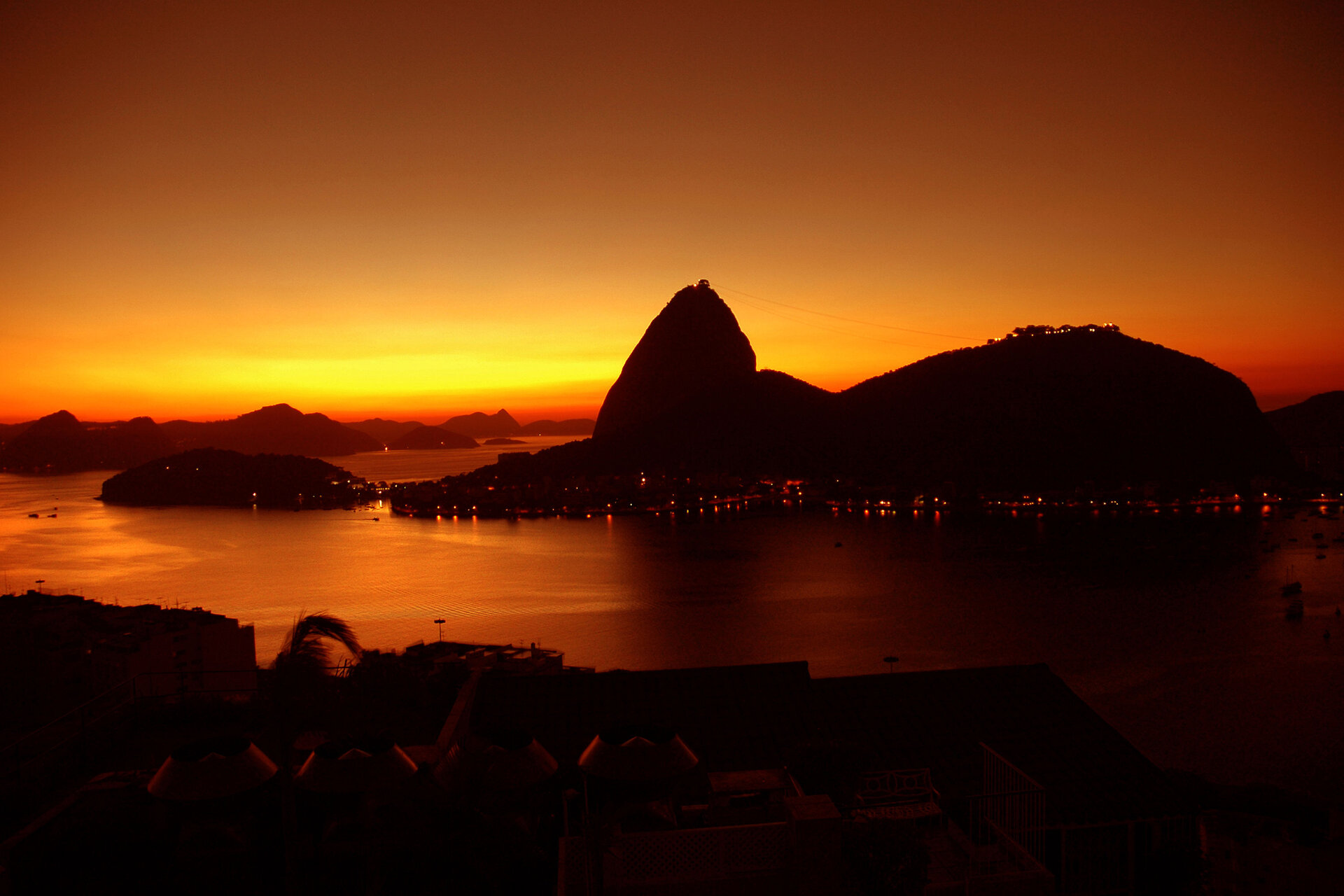 Spektakulärer Sonnenuntergang über dem Zuckerhut in Rio de Janeiro, Brasilien, mit einem leuchtend orangefarbenen Himmel und der Silhouette des Berges, der sich im ruhigen Wasser spiegelt.