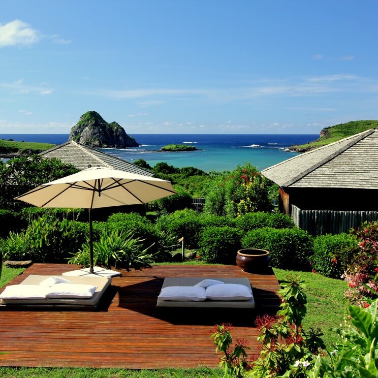 Entspannungsbereich im Garten der Pousada Maravilha in Fernando de Noronha, Brasilien, mit Liegen, Sonnenschirm und Blick auf das Meer.