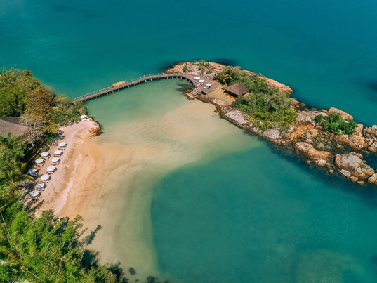 Luftaufnahme eines Strands mit Sonnenschirmen und Holzsteg zu einer kleinen Insel in Santa Catarina, Brasilien.