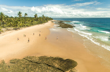 Perspektivische Ansicht der Praia dos Três Coqueiros auf Maraú. Im Vordergrund sind flache Felsformationen im Sand zu sehen. Eine Gruppe von Menschen spielt Fußball am weitläufigen Strand, der von unzähligen Kokospalmen gesäumt wird. Das Meer geht von hellem Türkis in ein tiefes Blau über, unter einem leicht bewölkten Himmel.