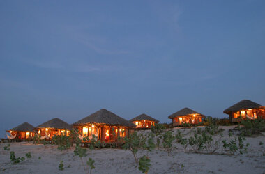 Reihe von beleuchteten Holzbungalows mit Reetdächern in der Dünenlandschaft bei Nacht.