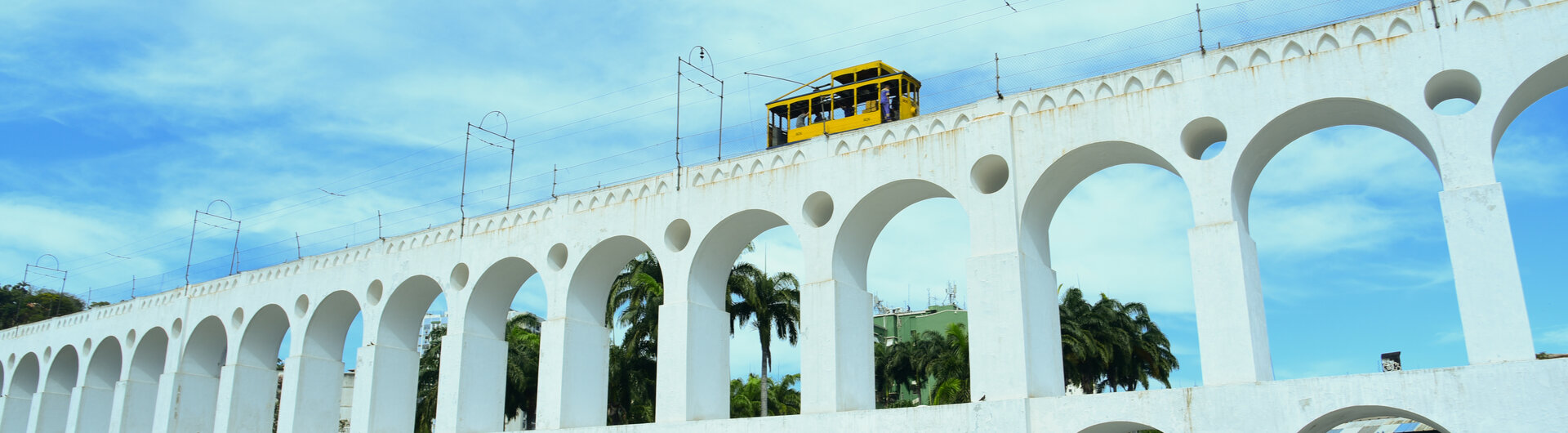 Das weisse Aquaedukt Arcos da Lapa in Rio de Janeiro mit der gelben Strassenbahn Santa Teresa unter blauem Himmel.