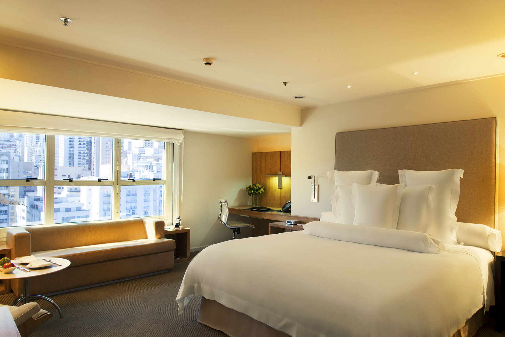 Zimmer im Hotel Emiliano in São Paulo mit einem komfortablen Bett, einem Sitzbereich und großen Fenstern mit Stadtblick.